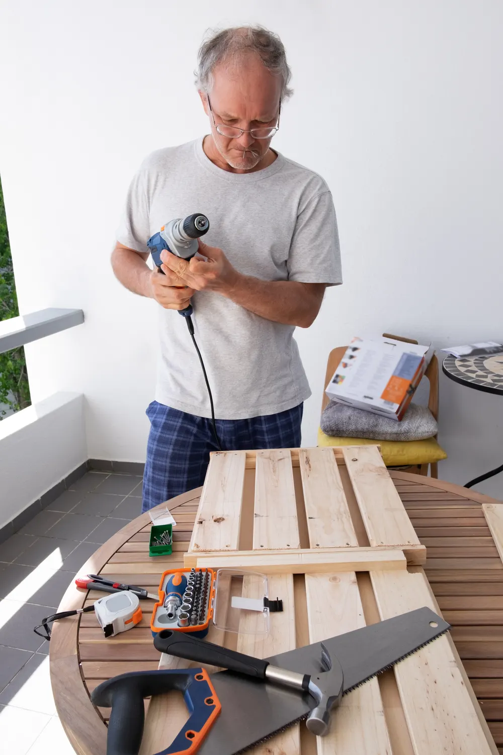 Heimwerker mit Bohrmaschine vor einem Tisch mit Brettern, Säge und Werkzeug