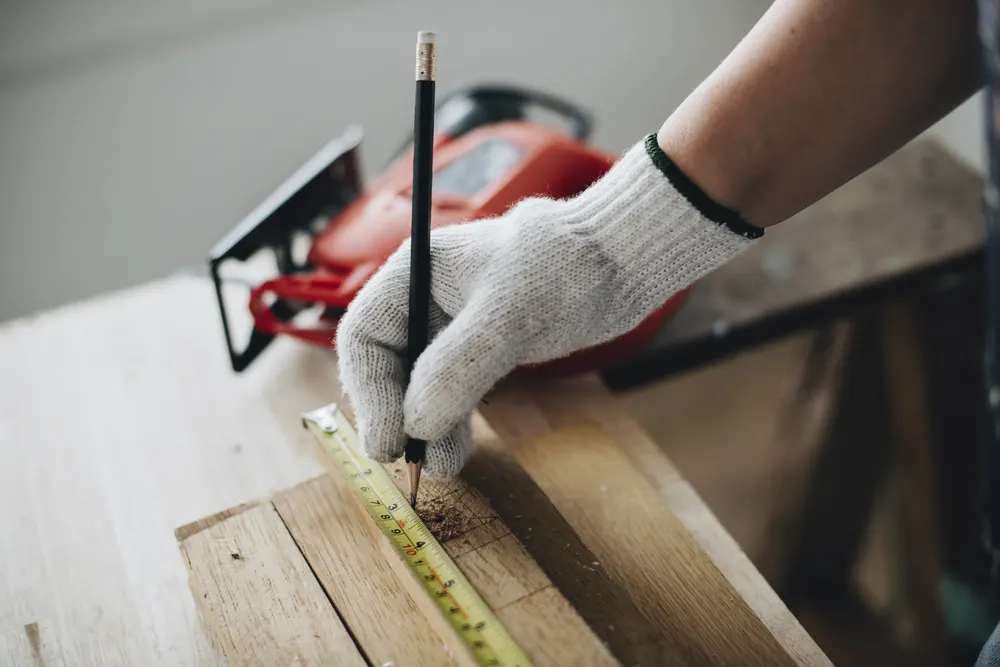 Hand im Arbeitshandschuh misst und markiert ein Holzbrett mit Bleistift und Maßband