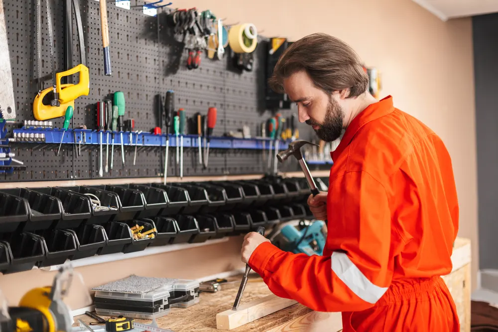 Handwerker in orangefarbener Arbeitskleidung arbeitet mit dem Hammer an einer Werkbank
